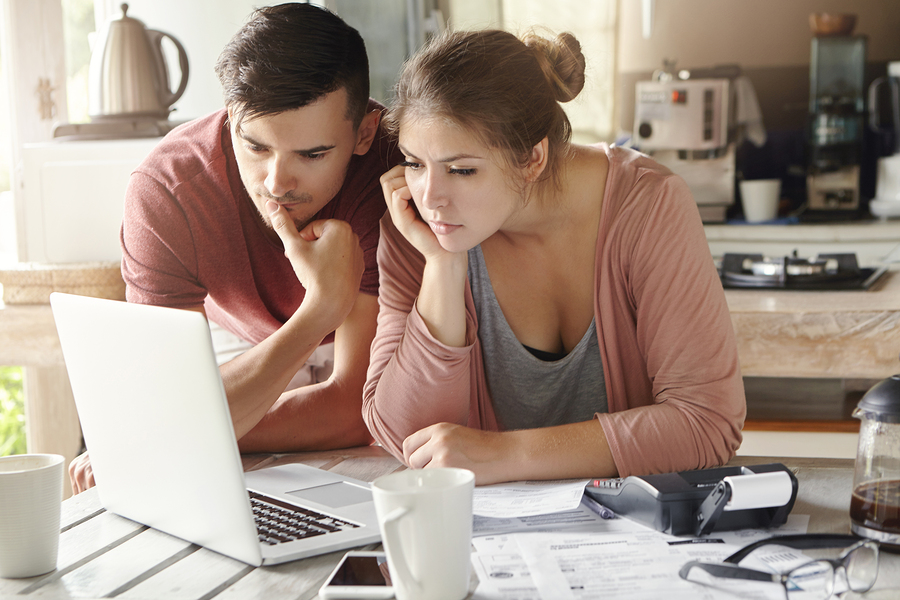 Serious man and woman sitting at kitchen table in front of open laptop computer looking at screen with concentrated expression focused on paying utility bills online. Family budget and finances